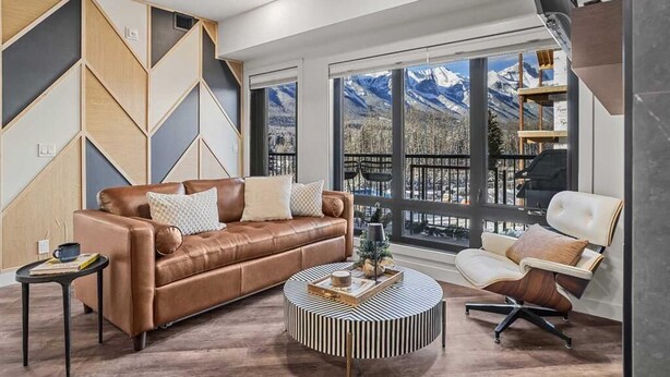 A modern living room with a brown leather sofa, a round coffee table, and a white armchair. Large windows offer a view of mountains. - New Luxury 3 bedroom (Canmore)