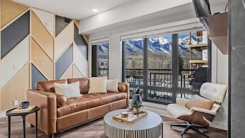A modern living room with a brown leather sofa, a round coffee table, and a white armchair. Large windows offer a view of mountains.