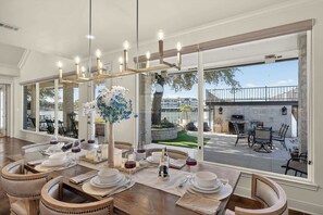 A dining area with a large table set for six, a chandelier, and a view of the outdoor patio. - Palm Cove | Concierge Services | Infinity Pool (Horseshoe Bay)