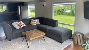 Una sala de estar moderna con un sofá seccional gris, una mesa de centro de madera y vistas a un jardín a través de grandes ventanales.