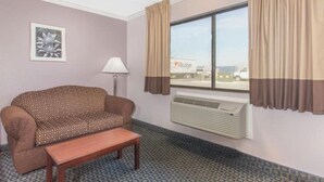 Ein Hotelzimmer mit einem gemusterten Sofa, einem kleinen Holztisch, einem Fenster mit Vorhängen, einer Wand-Klimaanlage und einem gerahmten Bild an der Wand.