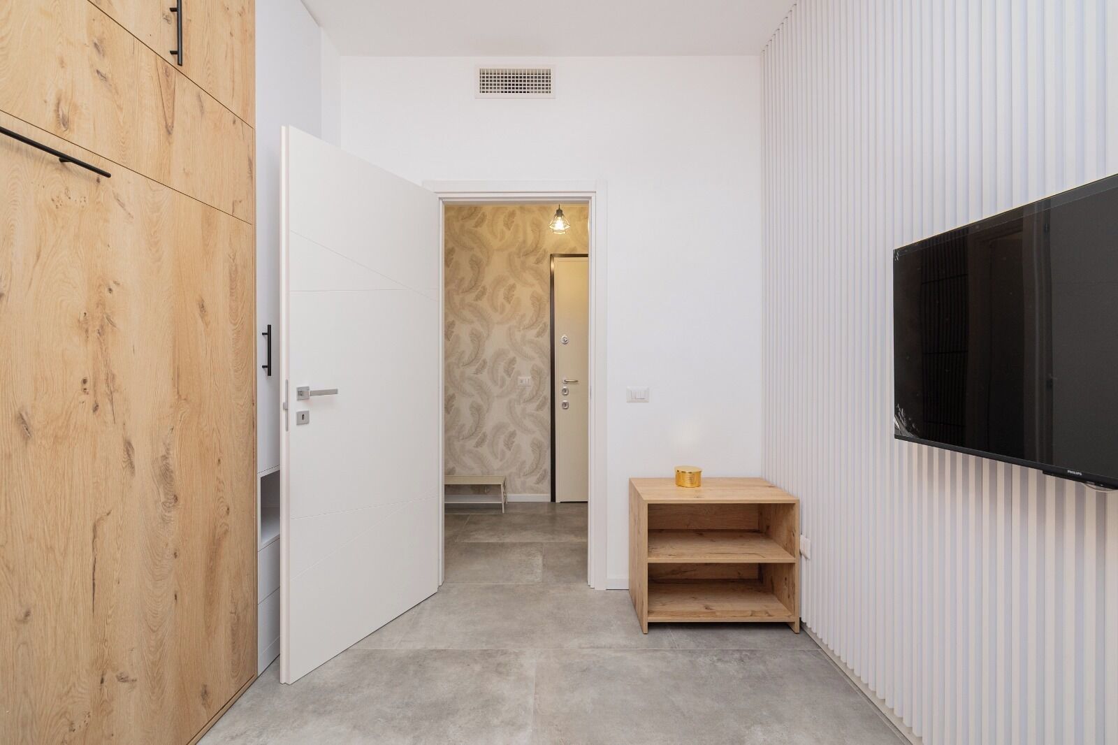 Een gang met een houten garderobe, een witte deur, een klein houten tafeltje en een tv aan de muur.