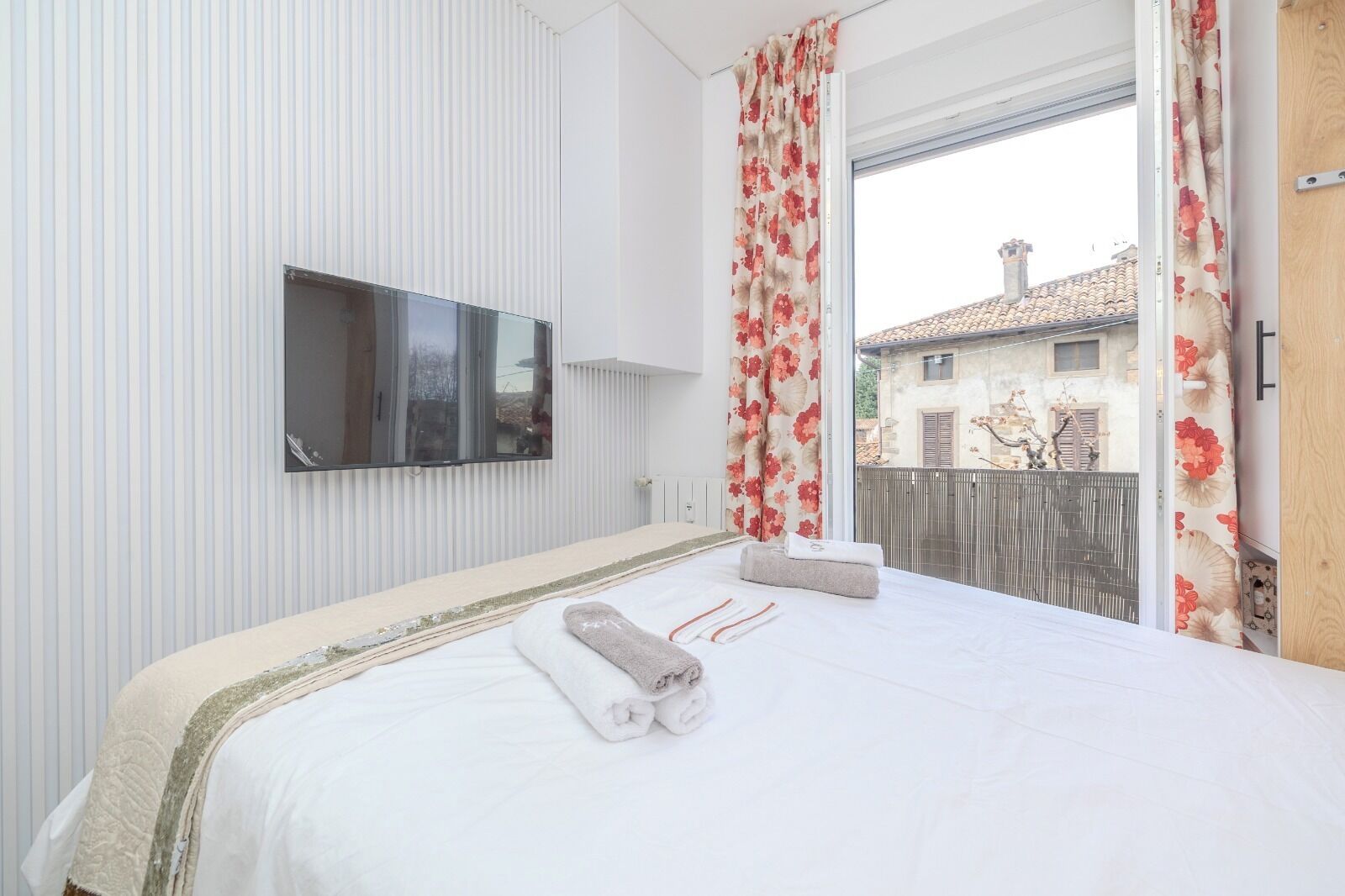 A hotel room with a large bed, a flat-screen TV mounted on the wall, and a balcony with a view of buildings and a fence.