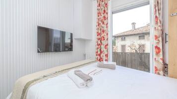 A hotel room with a large bed, a flat-screen TV mounted on the wall, and a balcony with a view of buildings and a fence.