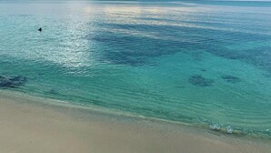 Ein Strand mit klarem türkisblauem Wasser und einer entfernten Bergkette unter bewölktem Himmel.