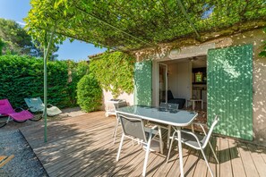 A wooden deck with a table and chairs, a covered seating area, and a building entrance with green shutters. - Charming Mazet in Secure Residence with Pool Near Alpilles and Town Center (Saint-Rémy-de-Provence)