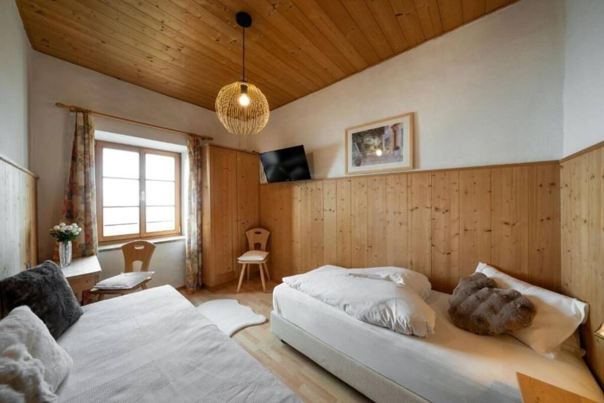 Une pièce aux murs recouverts de lambris en bois, avec un lit, une chaise, un bureau, un téléviseur et une fenêtre avec des rideaux.