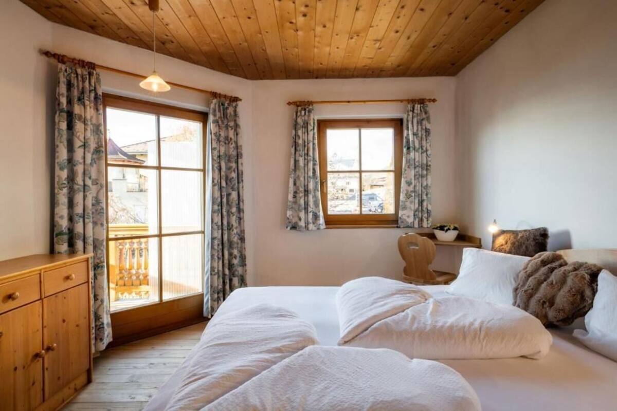 Une chambre avec un plafond en bois, un lit avec une literie blanche, deux fenêtres avec des rideaux et une commode en bois.