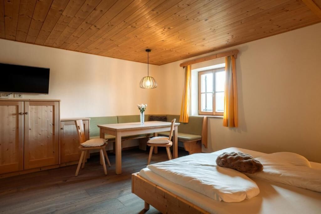 A room with a wooden ceiling, a bed, a desk with chairs, a television, and a window with curtains.