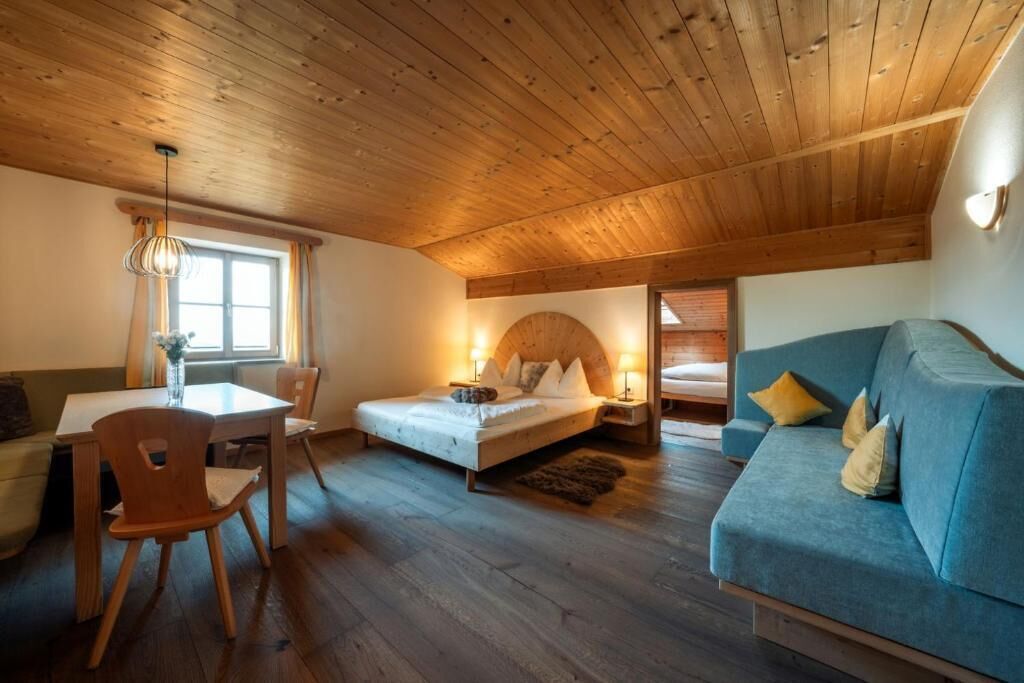 Een kamer met een houten plafond, een bed met kussens, een blauwe bank, een eettafel met stoelen en een raam met gordijnen.