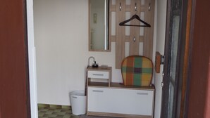 Une petite pièce, de dimensions réduites, avec un sol à carreaux, une armoire en bois dotée d’un miroir, un cintre noir suspendu et une petite poubelle.