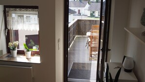 Une pièce avec un plancher en bois, une fenêtre ornée de rideaux en dentelle, un radiateur, une plante en pot et une porte donnant sur un balcon.