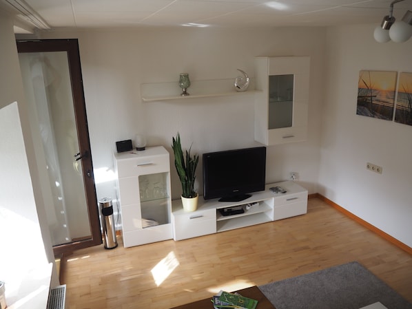 Un salon moderne avec un téléviseur à écran plat, des meubles blancs, une plante en pot et un plancher en bois.