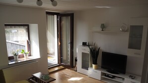 Un salon moderne avec un canapé, une table basse, un téléviseur posé sur un meuble et une porte-fenêtre coulissante donnant sur un balcon agrémenté de plantes en pot.