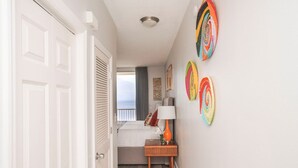 Un corridoio che conduce a una camera da letto con un letto, un comodino e una lampada. Sono presenti decorazioni murali colorate e una vista sull'oceano attraverso una finestra.