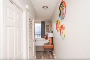 Ein Flur, der in ein Schlafzimmer mit Bett, Nachttisch und Lampe führt. Bunte Wanddekorationen und ein Blick auf das Meer durch ein Fenster sind zu sehen.