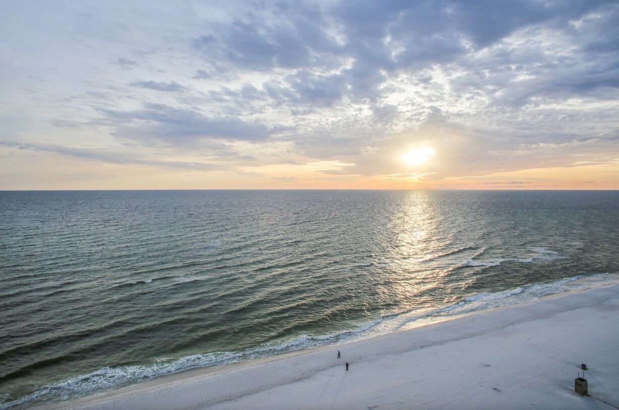 Une plage où l’on peut admirer le coucher de soleil sur l’océan, sous un ciel dégagé, et où quelques personnes se promènent sur le sable.