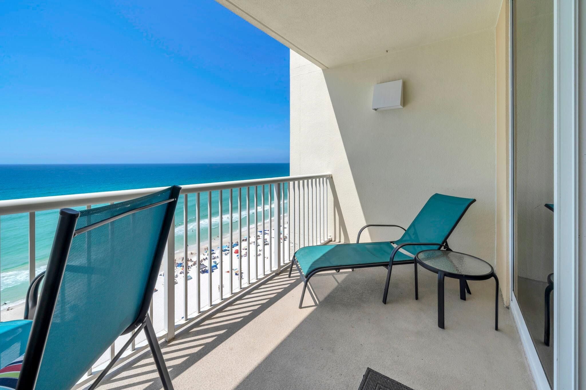 A balcony with a view of the beach and ocean, two green chairs, and a small table.