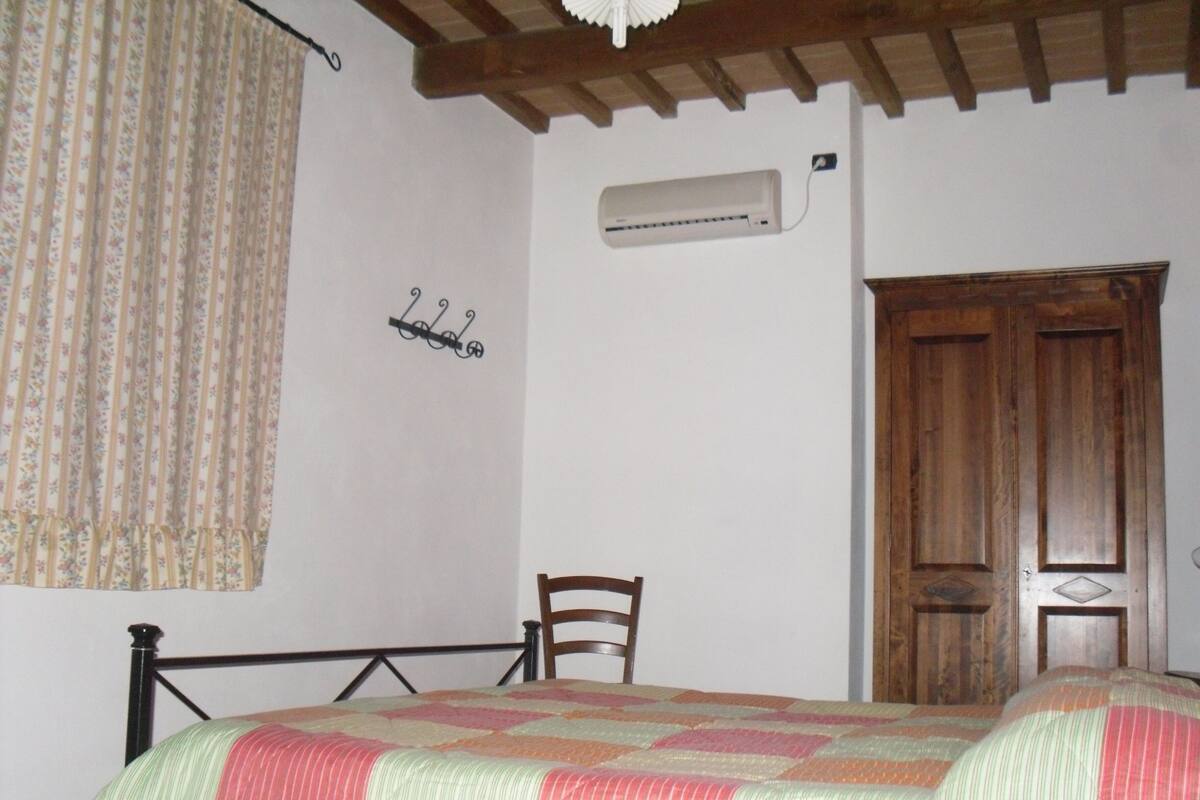 Un dormitorio con una cama, una silla, una puerta de madera y un equipo de aire acondicionado en la pared.