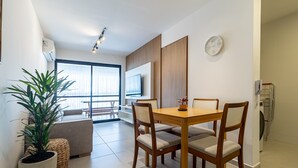 Ein modernes Wohnzimmer mit einem Holzesstisch, gepolsterten Stühlen, einer großen Topfpflanze und einem an der Wand befestigten Fernseher.