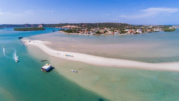 Luftaufnahme einer Küstenlandschaft mit Sandstrand, klarem türkisblauem Wasser und einer kleinen Insel mit Gebäuden.
