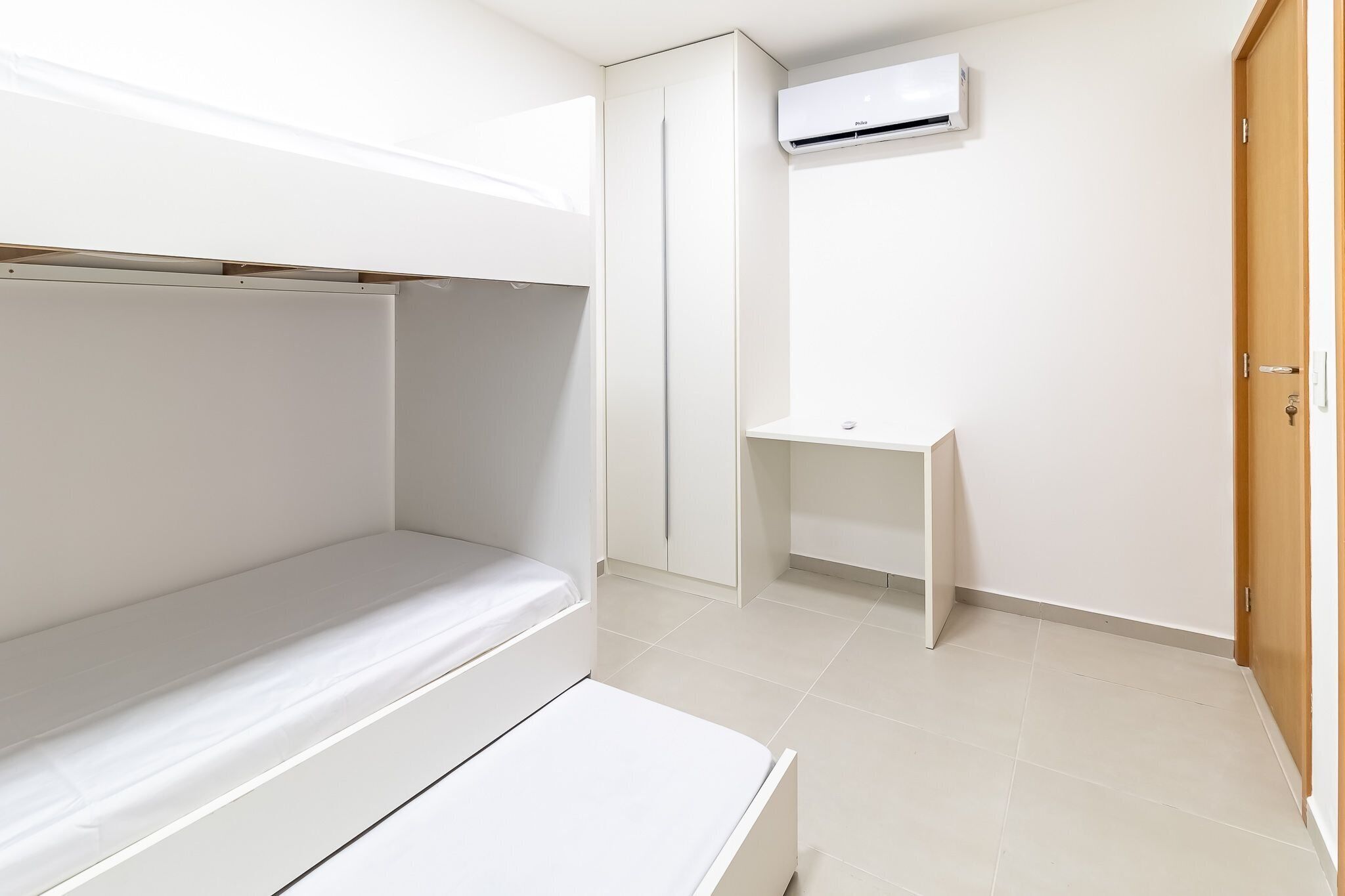 A small, white room with a bunk bed, a small desk, and an air conditioning unit on the wall.
