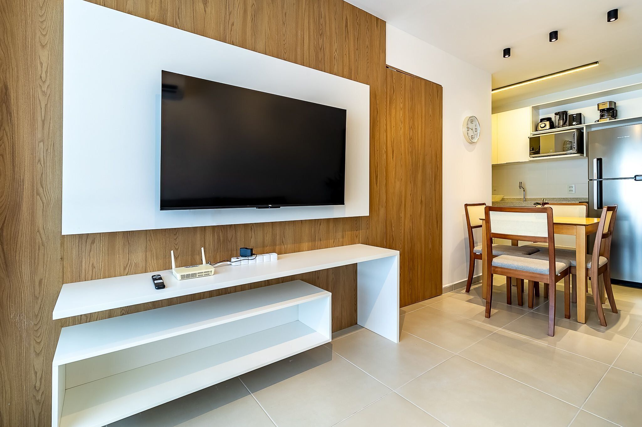 Uma sala de estar moderna com televisão montada, estantes brancas e um espaço de jantar com cadeiras e mesa de madeira.