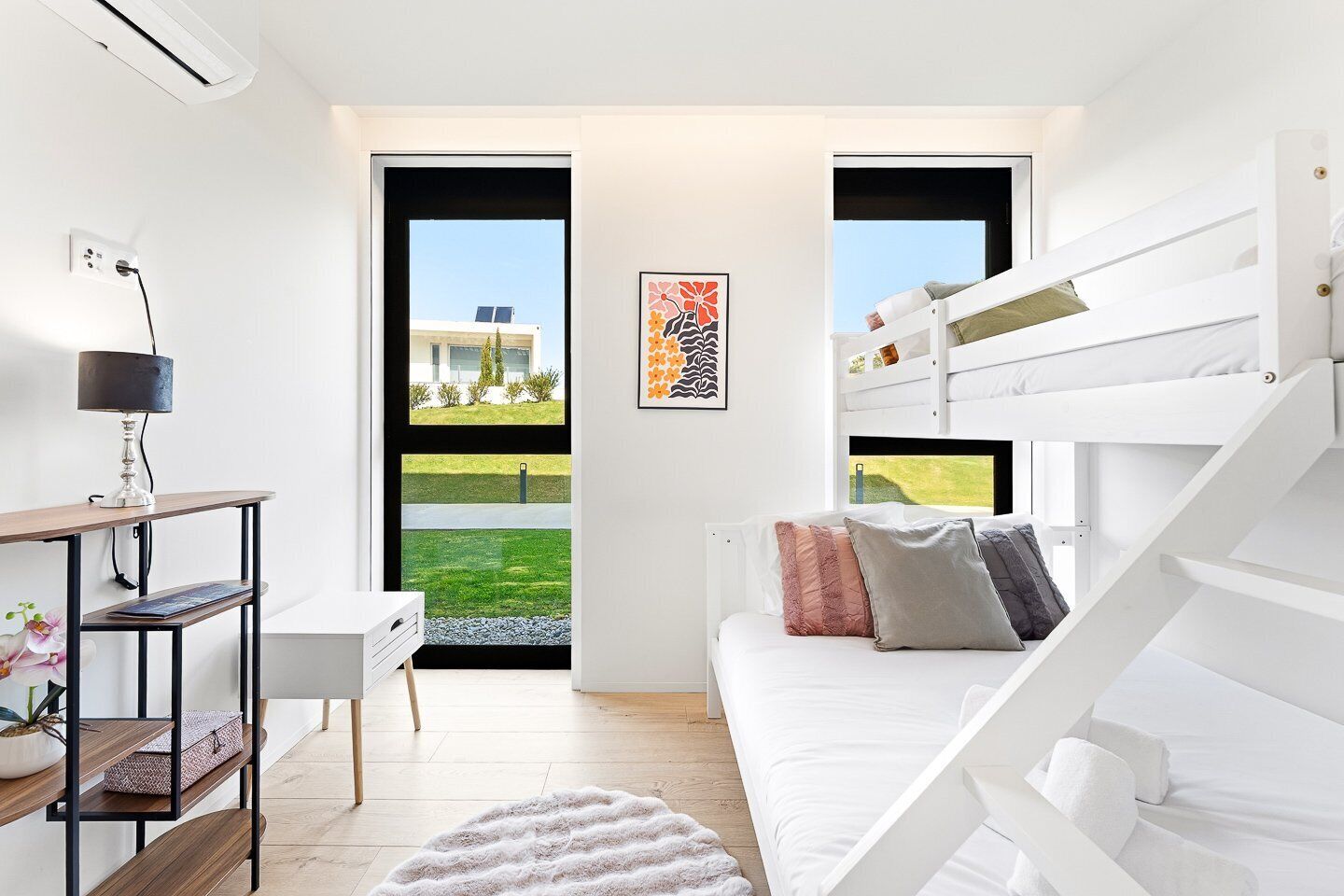 A modern bedroom with a bunk bed, a desk with a lamp, and a view of a building through a large window.