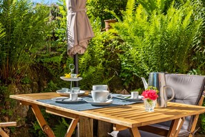 Ein Holztisch im Freien mit einem Stuhl, einer Metallkanne und einer Glasvase mit Blumen.