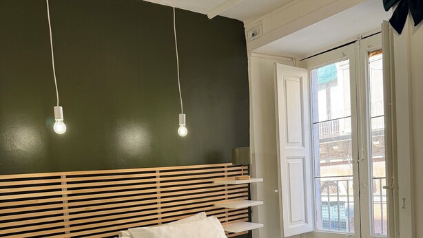 Un lit bien fait, avec une couette verte, une tête de lit en bois et des oreillers blancs. Deux lampes suspendues sont accrochées au plafond.