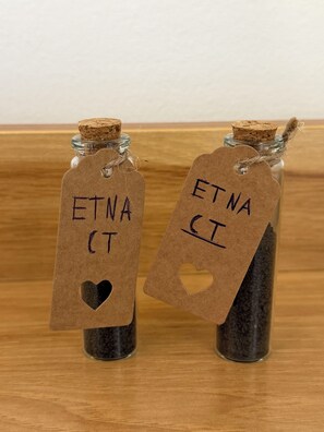 Zwei Glasflaschen mit Korken, jede mit einem Anhänger, auf dem „ETNA CI“ steht und ein Herz-Aussparung hat.