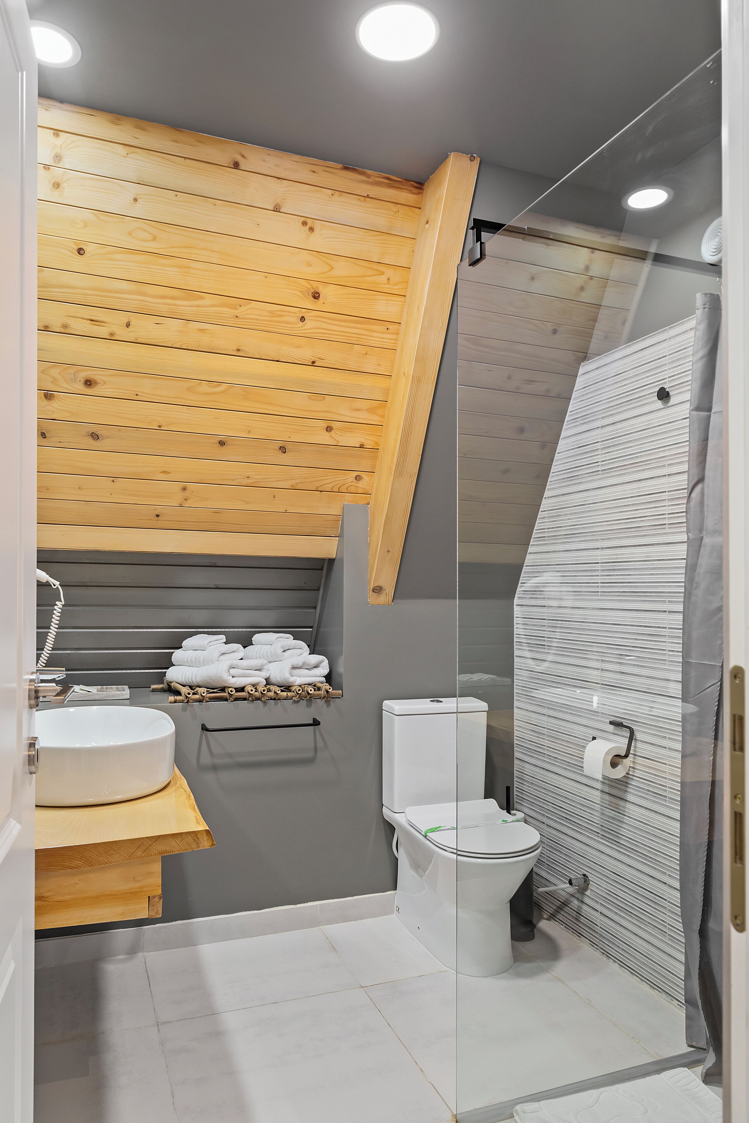 A modern bathroom with a wooden ceiling, a glass shower enclosure, a white sink, and a toilet.