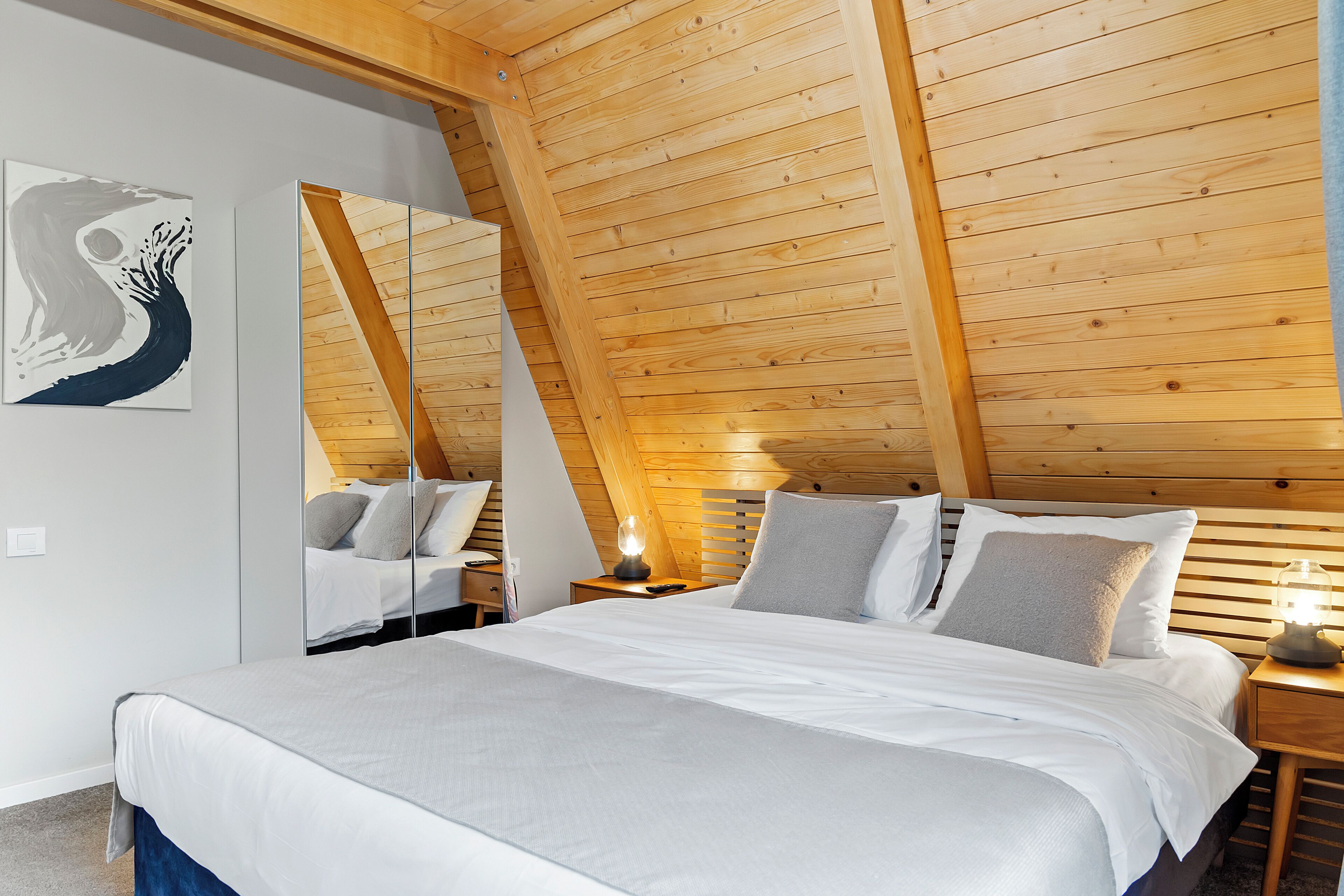 A bedroom with a wooden sloped ceiling, a bed with white bedding, a bedside table with a lamp, and a wall-mounted mirror reflecting the bed.