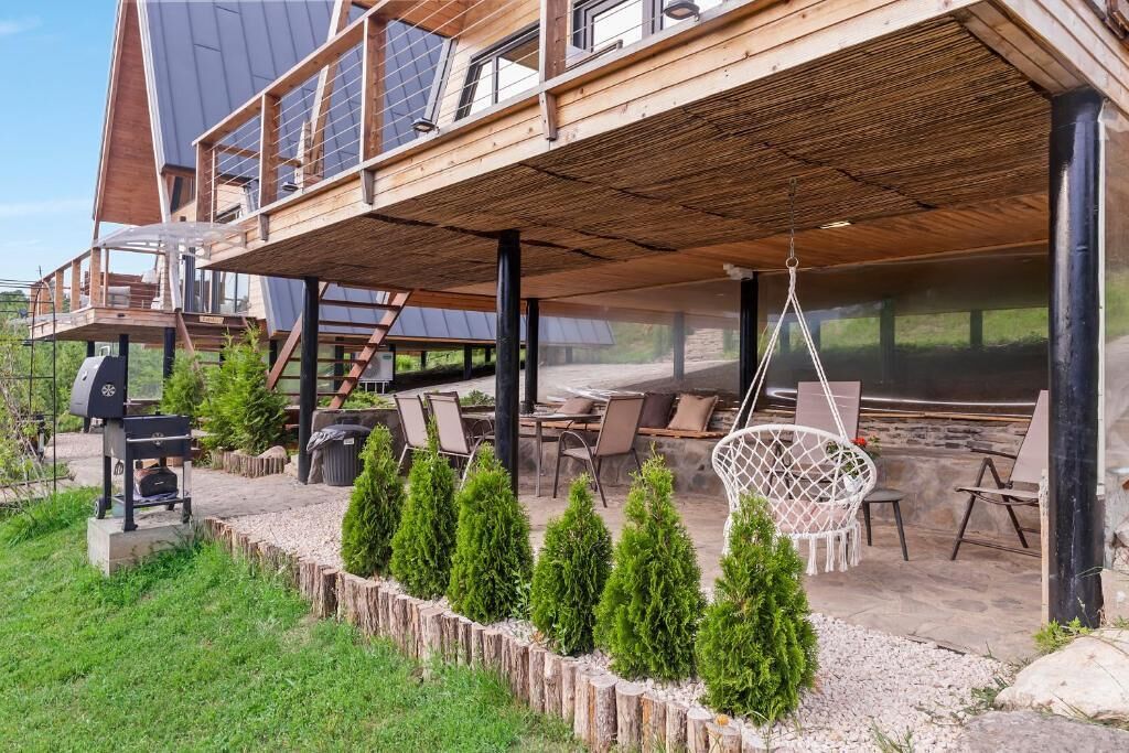 A modern wooden house with a covered patio area, outdoor seating, and a barbecue grill.