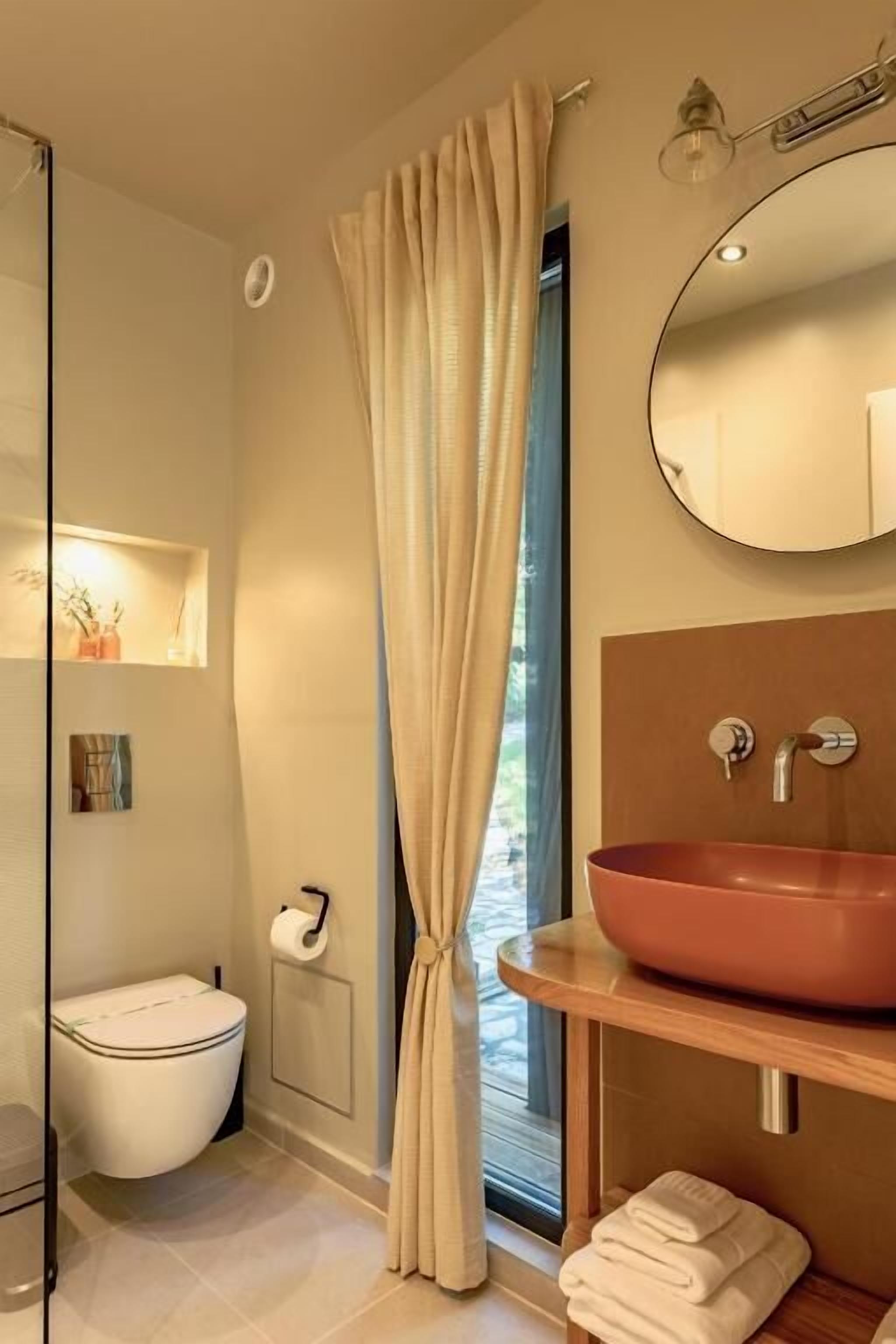 A modern bathroom with a glass shower, a beige toilet, a wooden vanity with a red basin, and a large mirror.