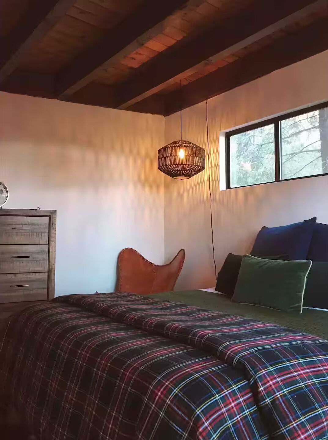 Ein Schlafzimmer mit karioter Bettdecke, einem Holzkleiderschrank, einer gewobenen Leuchte und einem Fenster mit Blick auf Bäume.