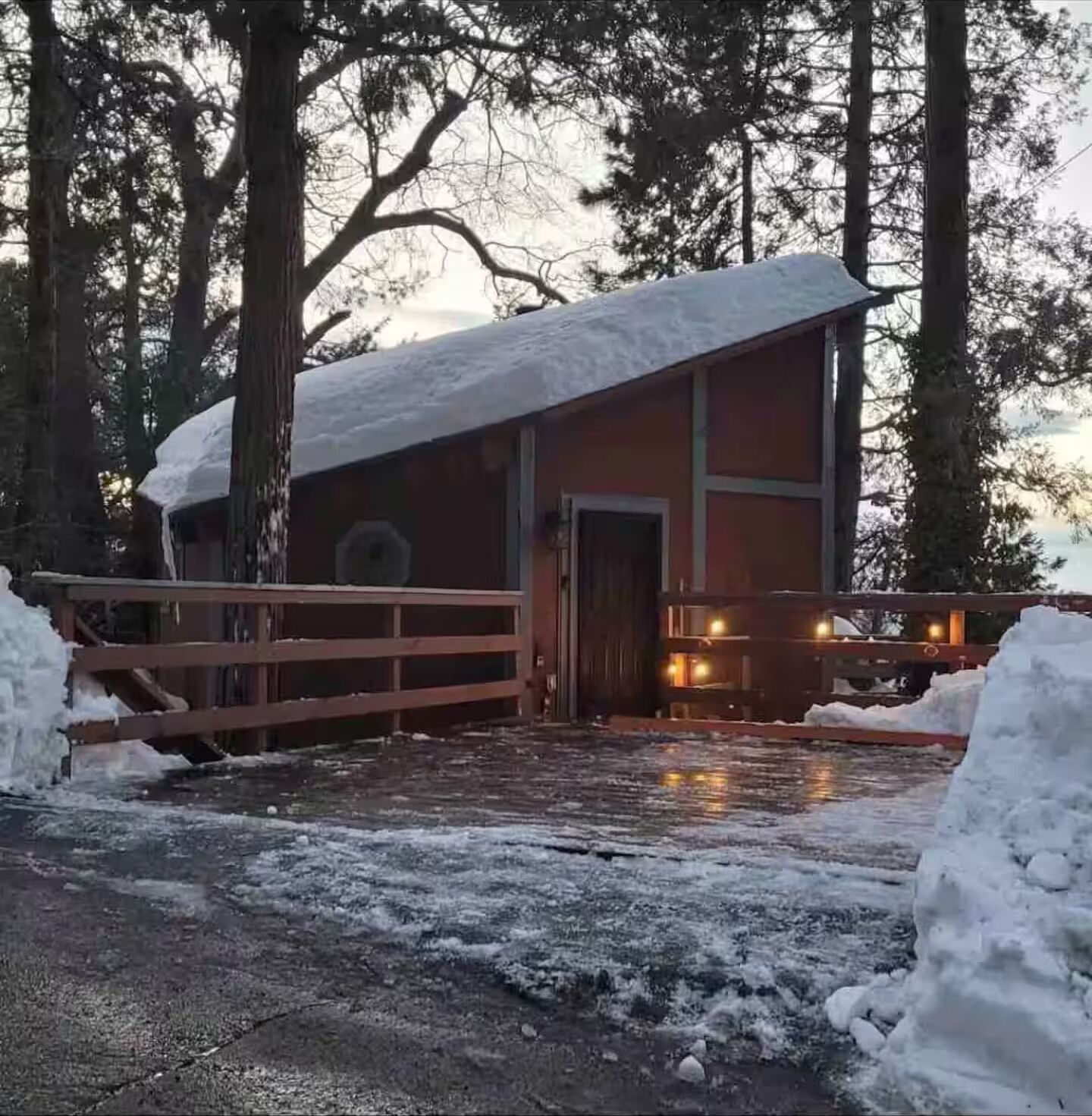 Eine Holzhütte mit einer Holzterrasse und einem mit Schnee bedeckten Dach, umgeben von hohen Bäumen.