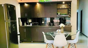 Una cocina moderna con armarios oscuros, un refrigerador de acero inoxidable, una mesa de comedor de vidrio con sillas blancas y un jarrón con flores sobre la mesa.