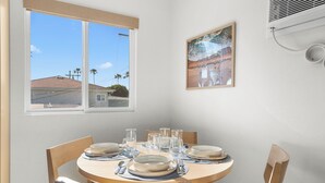 Un coin repas avec une table ronde pour quatre personnes, des chaises, une fenêtre donnant sur des immeubles et un appareil de climatisation.