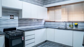 Una cocina con gabinetes blancos, una estufa negra, encimera de mármol, ventana con persianas y un lavamanos con productos de limpieza.