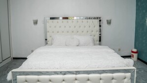 Una cama impecablemente vestida, con un cabecero capitonado, una colcha blanca y esponjosa y un piecero de mármol.