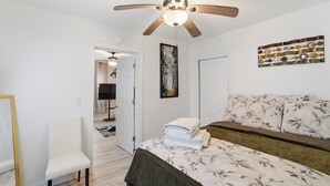Un dormitorio con cama, una silla, ventilador de techo y una obra de arte en la pared.