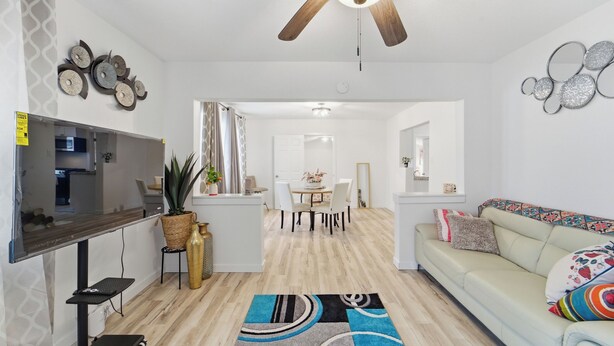 Una sala de estar moderna con una televisión de pantalla plana, un ventilador de techo, una zona de comedor con una mesa y sillas, y un sofá con cojines.