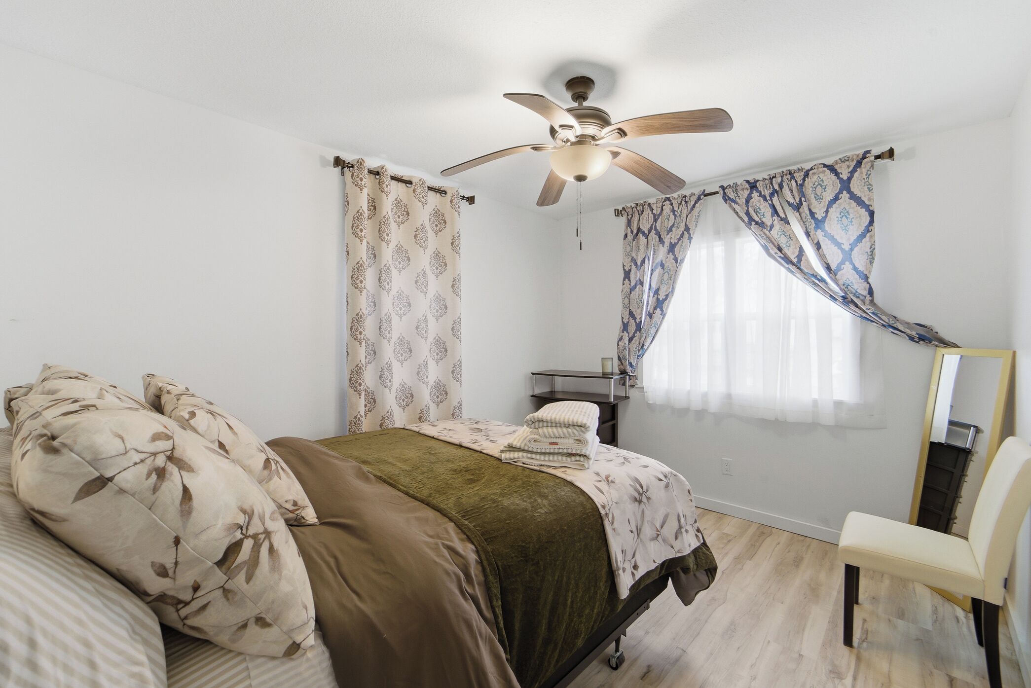 Ein Schlafzimmer mit Bett, Deckenventilator, Stuhl und einem fenster mit Vorhängen.