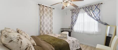 Ein Schlafzimmer mit Bett, Deckenventilator, Stuhl und einem fenster mit Vorhängen.