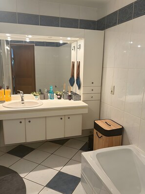 Ein Badezimmer mit einem weißen Waschbecken, einem Spiegel und einer Badewanne. Auf dem Waschbecken stehen Pflegeprodukte, es gibt einen Handtuchhalter und einen Mülleimer.