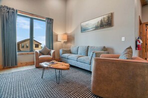 Ein Wohnzimmer mit Sofa, Sessel und Couchtisch, einem Gemälde an der Wand und einem Fenster mit Vorhängen.