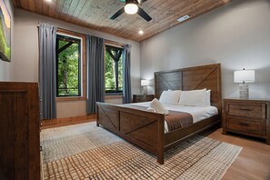 A bedroom with a wooden bed, a nightstand, a lamp, and a view of greenery through the windows. - 5BR Luxury Cabin・Pool・Near Dollywood / Water Parks (Sevierville)