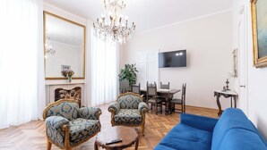 Ein Wohnzimmer mit einem blauen Sofa, zwei gemusterten Sesseln, einem Holztisch, einer Chandelier, einem Fernseher und einem Essbereich mit Tisch und Stühlen.