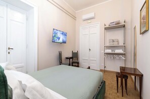 Ett sovrum med en säng, en tv, en stol, ett litet bord och en hylla med galgar.